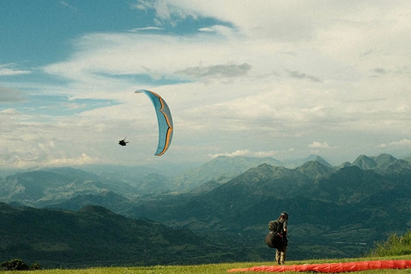 Two people preparing for paragliding in Jericó, Colombia, one flying above the mountains, one getting ready on the ground.